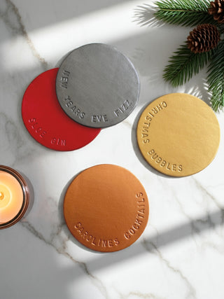 Four round coasters with embossed text on a marble surface with a candle and pine branches.