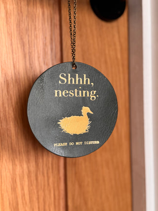 Olive green leather door hanger that says shh nesting on it in gold with a gold duck. Under the duck it reads 'please do not disturb'.