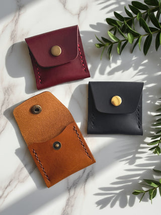 Three leather pouches in red, navy, and tan brown on a marble surface with green leaves.