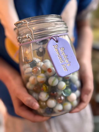 A young boy holding a marble jar wrapped with a hand-painted lavender purple label that reads "Sophie's Reward Jar" in neat, handwritten-style lettering. The label adds a personal and cheerful touch to the jar, which is likely used for collecting rewards or incentivess