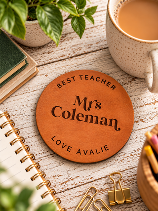 A round tan leather coaster engraved with 'BEST TEACHER Mr's Coleman LOVE AVALIE' in black lettering, styled on a white wood tabletop with a mug of coffee, notebook, binder clips, and plants.