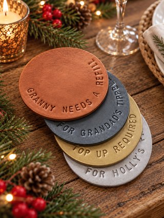 Four round castor-style coasters stacked on a rustic wooden table, each embossed with a humorous holiday phrase. The coasters are shown in copper, dark gray, gold, and light gray, surrounded by festive holiday decorations including pine branches, red berries, a lit candle, and a wine glass.