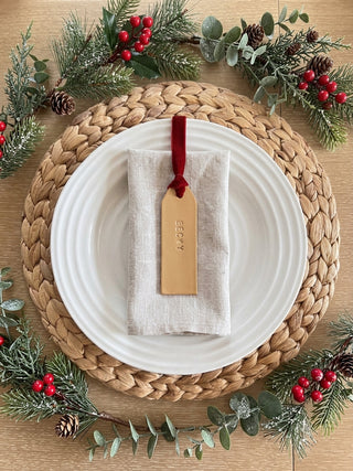 A styled holiday table setting featuring a white ceramic plate on a woven straw placemat, with a folded beige linen napkin and a tan leather gift tag tied with a red ribbon. The scene is decorated with green eucalyptus, pine branches, and red berries on a wooden tabletop.