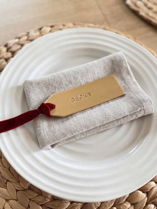 A personalised beige linen gift pouch with a red velvet ribbon and a gold metal tag engraved with the name 'DEOIL', displayed on a white ceramic plate over a woven placemat.