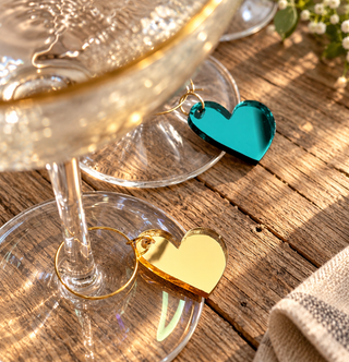 Two small heart-shaped wine tag charms, one gold and one teal, attached to the bases of wine glasses on a rustic wooden table.