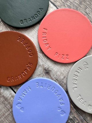 Hand-stamped leather coasters in various colours, featuring personalised embossed text such as 'Grandpa's Coffee,' 'Friday Fizz,' and 'Herbal Tea.' Perfect for custom gifting and home decor