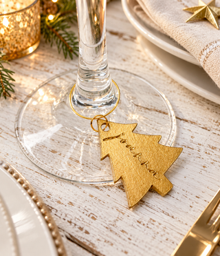 A gold leather Christmas tree-shaped ring tag engraved with 'Love, Bella' hangs from a wine glass stem on a rustic white wooden table with holiday decorations.