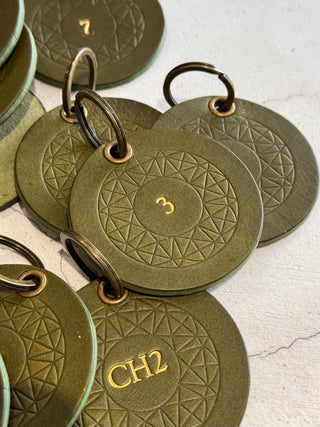 A close-up of olive green circular key tags with embossed geometric patterns and brass key rings, some showing gold-colored numbers and letters like '3' and 'CH2'.