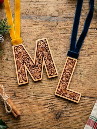 Two wooden monogram letter ornaments, 'M' and 'L', with intricate laser-cut floral patterns, hanging from velvet ribbons (one yellow, one navy blue) on a rustic wooden tabletop.
