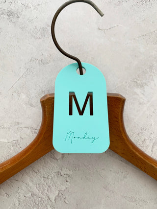 A blue acrylic hanger tag with the letter 'M' for Monday attached to a wooden hanger.
