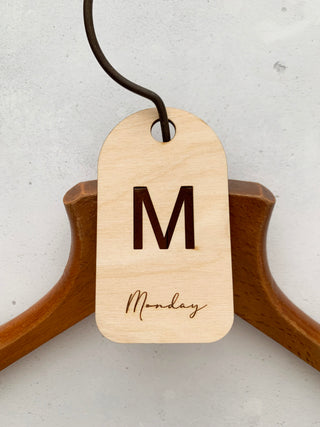 A wooden hanger tag with the letter 'M' and the word 'Monday' engraved on it, hanging on a brown wooden hanger.