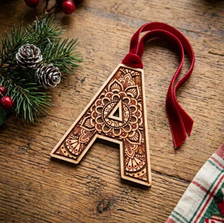 A wooden letter A Christmas ornament with intricate laser-engraved mandala patterns and a red velvet hanging ribbon, displayed on a rustic wooden tabletop with pine branches and holiday decorations.