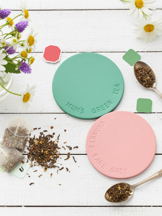Two leather coasters one seafoam green and one pink, on a white wooden surface with tea leaves and teabags.