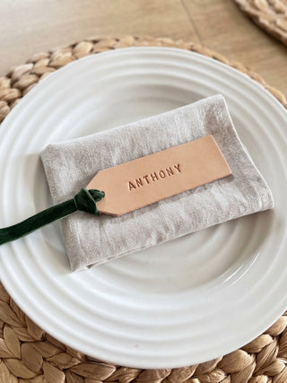 A personalized linen napkin with a tan leather tag engraved with the name 'ANTHONY' and a dark green ribbon, displayed on a white plate over a woven placemat.