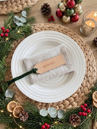 A styled holiday table setting featuring a white plate on a woven placemat, with a folded beige napkin and a personalized leather name tag that reads 'ANTHONY'. The setting is decorated with a pine wreath with fairy lights, red berries, pinecones, dried orange slices, and a small Christmas tree ornament on a wooden table.