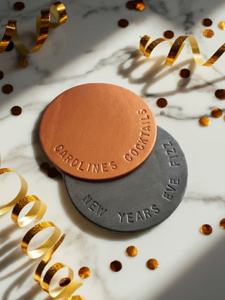 Two round coasters with 'Caroline's Cocktails' branding on a marble surface with gold confetti.