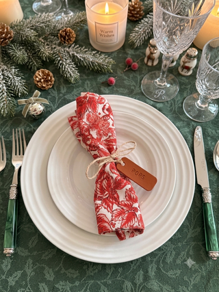 A festive holiday table setting featuring a white ceramic charger plate with a rolled napkin in a red and white floral pattern tied with twine and a small wooden tag. The setting is decorated with green pine branches, pinecones, a lit candle, and elegant glassware on a dark green damask tablecloth.