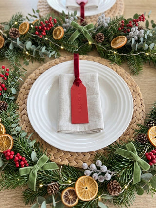 A festive holiday table setting featuring a white ceramic plate on a woven straw placemat, folded in a light beige napkin with a red fabric tag hanging from a red ribbon. The setting is surrounded by a lush green garland decorated with dried orange slices, red berries, pinecones, and green bows.
