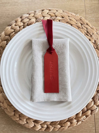 A folded light beige linen napkin with a red velvet ribbon and matching red tag attached, placed on a white plate over a woven placemat on a wood surface.