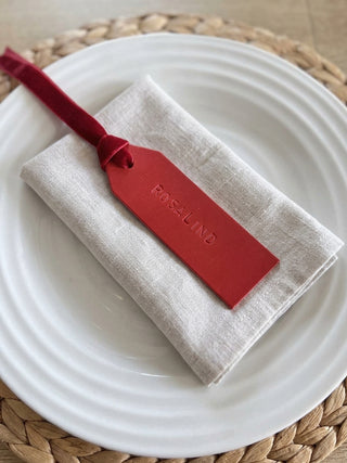 A folded light beige linen napkin with a red velvet ribbon and a matching red leather tag embossed with the brand name 'ROSALIND', presented on a white plate over a woven placemat.