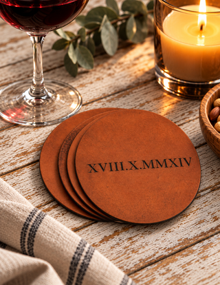 Rustic tan leather coasters with roman numbers on, lying on a rustic wooden table with a lit candle and glass of red wine.