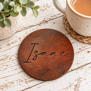 A round brown wooden coaster engraved with the name 'Isaac' in black script, styled on a white distressed wooden table next to a mug and a potted plant.