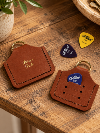 Two tan leather guitar pick holders with brass key rings on a rustic wooden table, with three guitar picks placed nearby.