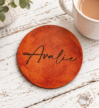 A round wooden coaster with the name 'Avalie' written in black script, placed on a distressed white wooden table next to a white mug and a small plant.