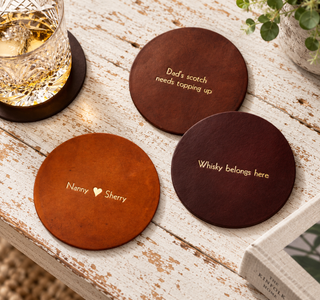 Three round wooden whiskey coasters in different wood tones, each engraved with a humorous or sentimental phrase, arranged on a distressed white wooden table next to a glass of whiskey.