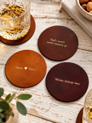 Three personalized leather coaster drink mats in brown shades with gold foil text, arranged on a rustic white wooden table alongside a glass of whiskey and a small plant.