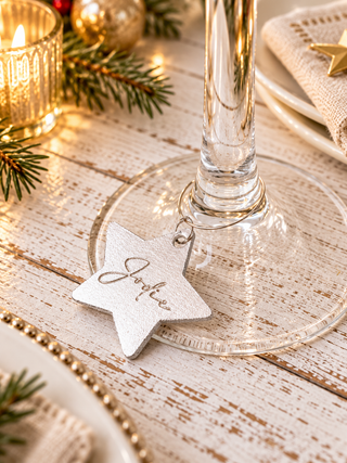 A silver leather glittery star-shaped wine glass charm engraved with the name 'Julie', hanging from the base of a clear glass wine glass on a rustic white wooden table decorated for Christmas with pine branches, a candle, and a napkin.