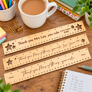 Three personalized wooden ruler bookmarks are arranged diagonally on a wooden desk, surrounded by office props like a white coffee mug, binder clips, sticky notes, and a notebook.