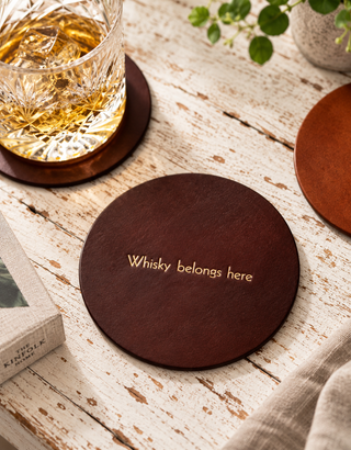 A round dark brown wooden coaster with gold foil text reading 'Whisky belongs here' sits on a distressed white wooden table next to a glass of whiskey and other coasters.