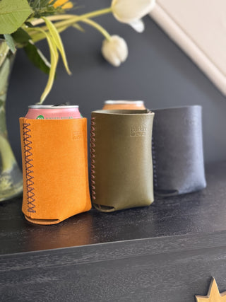 Three leather can coolers Koozie in yellow, green, and navy on a dark surface with flowers in the background.
