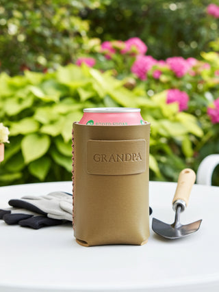 Olive Green can holder with 'GRANDPA' on it, containing a pink can, placed on a table outdoors with flowers and gardening tools in the background.