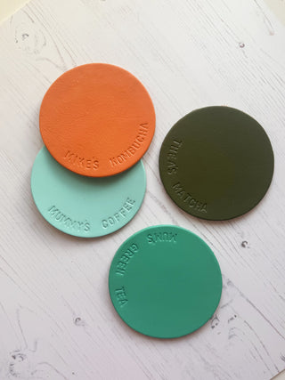 Four round coasters in orange, green, and teal on a textured white surface.