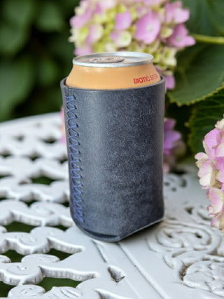 Navy blue leather handmade can cooler with a can of Bionic Soda on a decorative metal table with pink flowers in the background.