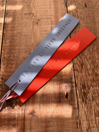 Two handcrafted leather bookmarks in coral and purple each embossed with sentimental messages: 'LOVE YOU MUMMY,' and 'TO MANY CHAPTERS TOGETHER.' Each bookmark has a matching leather tassel and is displayed on a rustic wooden surface with soft natural lighting