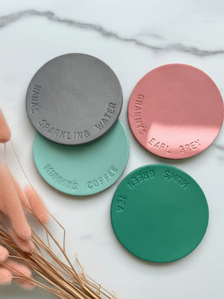 Four round coasters in soft pastel different colours with text on a marble surface.