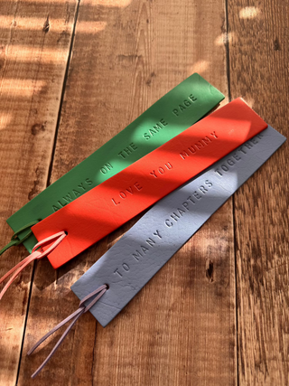 Three handcrafted leather bookmarks in green, coral, and grey, each embossed with sentimental messages: 'ALWAYS ON THE SAME PAGE,' 'LOVE YOU MUMMY,' and 'TO MANY CHAPTERS TOGETHER.' Each bookmark has a matching leather tassel.