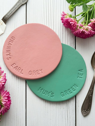 Two round coasters, one pink and one green, with engraved text on a wooden surface with flowers and cutlery.