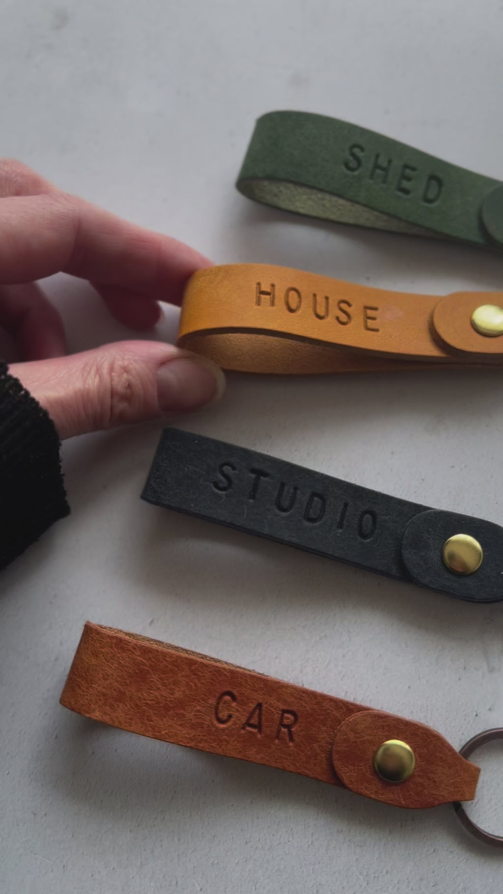 Handmade personalised leather keyrings by Parkin & Lewis, featuring embossed labels for car, house, studio, and shed keys, in orange, yellow, olive green, and navy leather with brass and bronze hardware