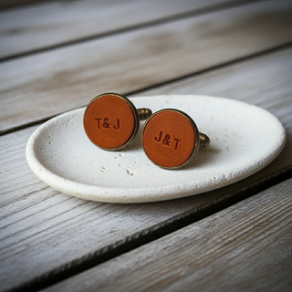 Pair of tan leather cufflinks with 'T&J' engraving on a white dish, placed on a wooden surface.