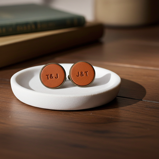 Tan Leather Brown cufflinks with 'T&J' engraving on a white dish on a wooden surface.